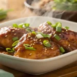 Close-up of glazed teriyaki chicken thighs in a white bowl, garnished with scallions and sesame seeds.